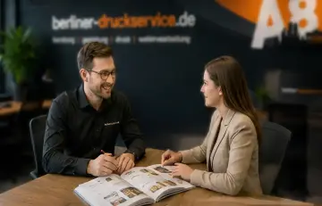 Ein Mann mit Brille und eine Frau in einem beigen Sakko sitzen sich an einem Holztisch gegenüber, lächeln und schauen gemeinsam in ein aufgeschlagenes Buch mit Produktbeispielen.