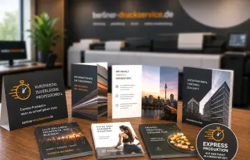 Mehrere aufgestellte und liegende Informationsbroschüren und Flyer mit unterschiedlichen Cover-Motiven auf einer Holzoberfläche. Im Hintergrund stehen große, dunkle Druckmaschinen.