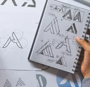 Typesetting and layout: Hand holding sketchpad with logo and design drafts on the desk.